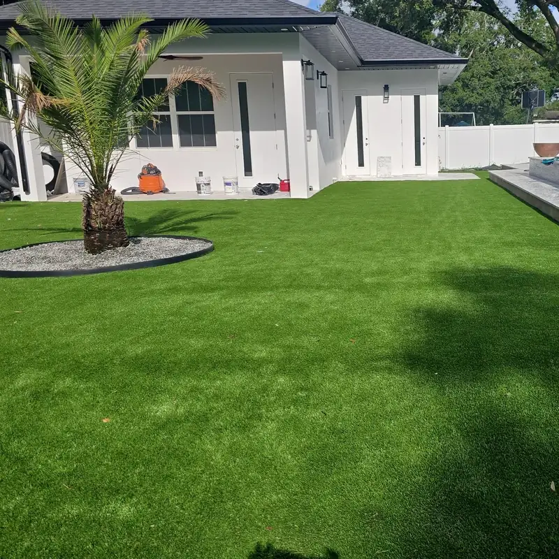 Artificial grass backyard at a white single-story home, with a palm inside a gravel ring and the pool deck at the far edge
