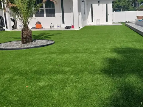 Artificial grass backyard at a white single-story home, with a palm inside a gravel ring and the pool deck at the far edge