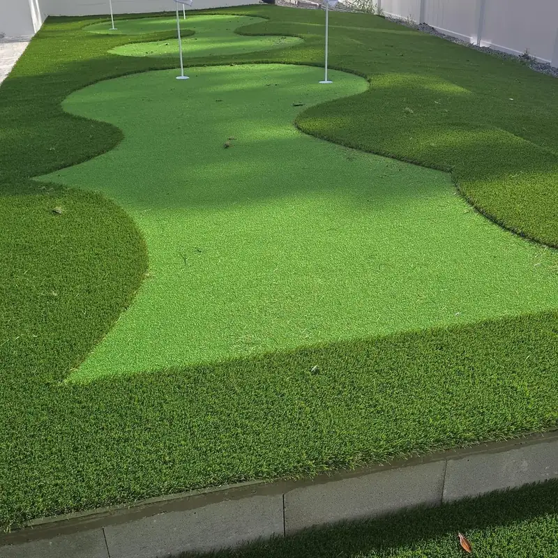 Backyard artificial putting green with three cups and flags, a taller fringe collar and a white vinyl privacy fence