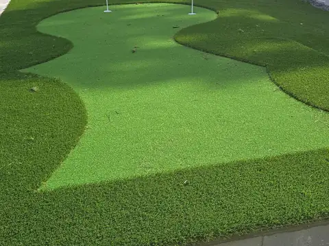 Backyard artificial putting green with three cups and flags, a taller fringe collar and a white vinyl privacy fence
