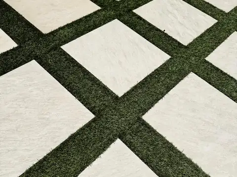 Close-up of artificial turf strips between square porcelain pavers set on the diagonal, with a block retaining wall behind