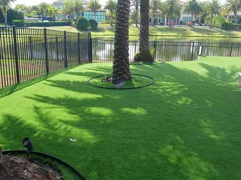 Lakefront backyard with artificial turf, two palms inside black ring edging, an aluminum picket fence and homes across the water